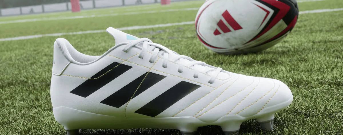 White adidas rugby boot beside a rugby ball on grass